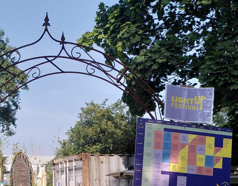 Blauer Himmel, Bäume und ein Weg mit einem Metallbogen, an dem Infos zum LightUp Bildungsfestival hängen.