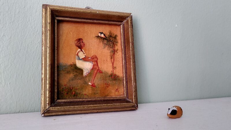 ein quadratische Bild eines Mädchens, das unter einem Baum sitzt und Vögel darauf anschaut, simpel gemalt auf Holz mit Rahmen. Davor auf einem Sims ein mini-kleines Keramik-Meerschwein, dem Bild zugewandt.